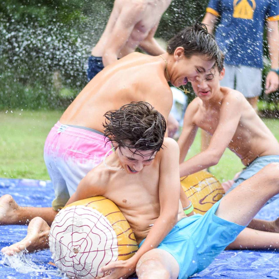 legacy campers wrestle over inflatable log while getting sprayed with water during summer camp