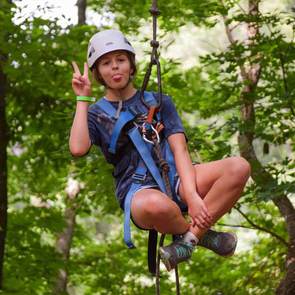 female legacy camper gives a peace sign with legs crossed while hanging in her zipline harness