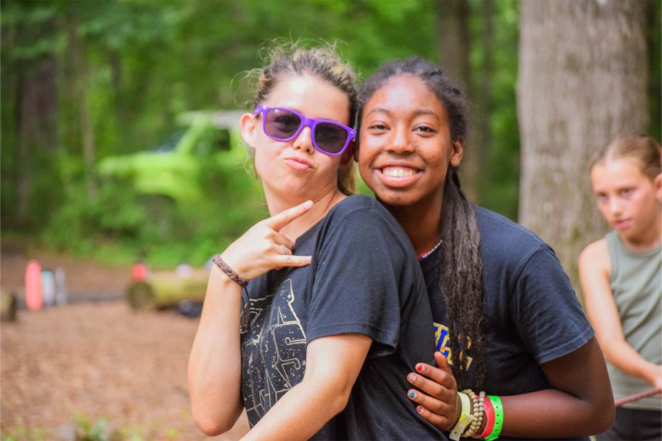 two female counselors, one wearing purple sunglasses embrace and pose for camera during legacy camp