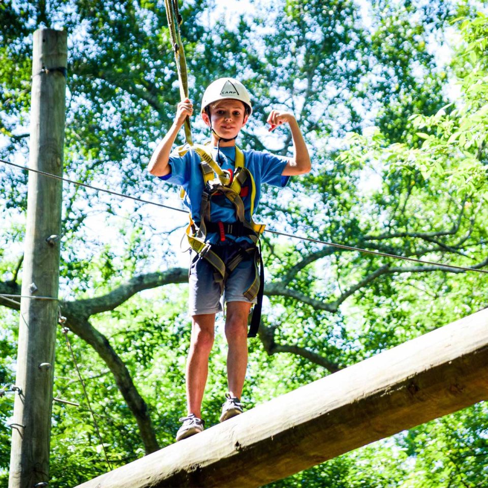 young male camper in white helmet and harness walks across high ropes course element at Camp Canaan