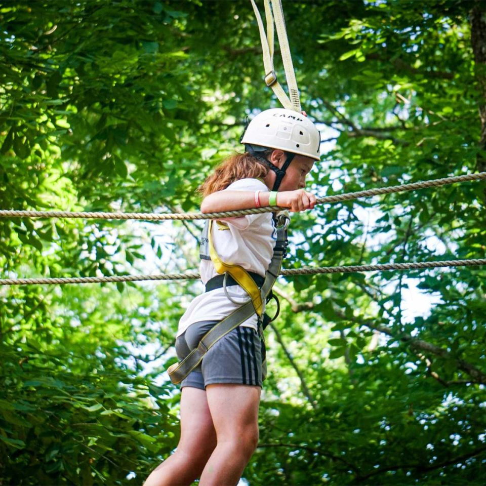 girl camper holds on to ropes while crossing high ropes course element boy at Camp Canaan