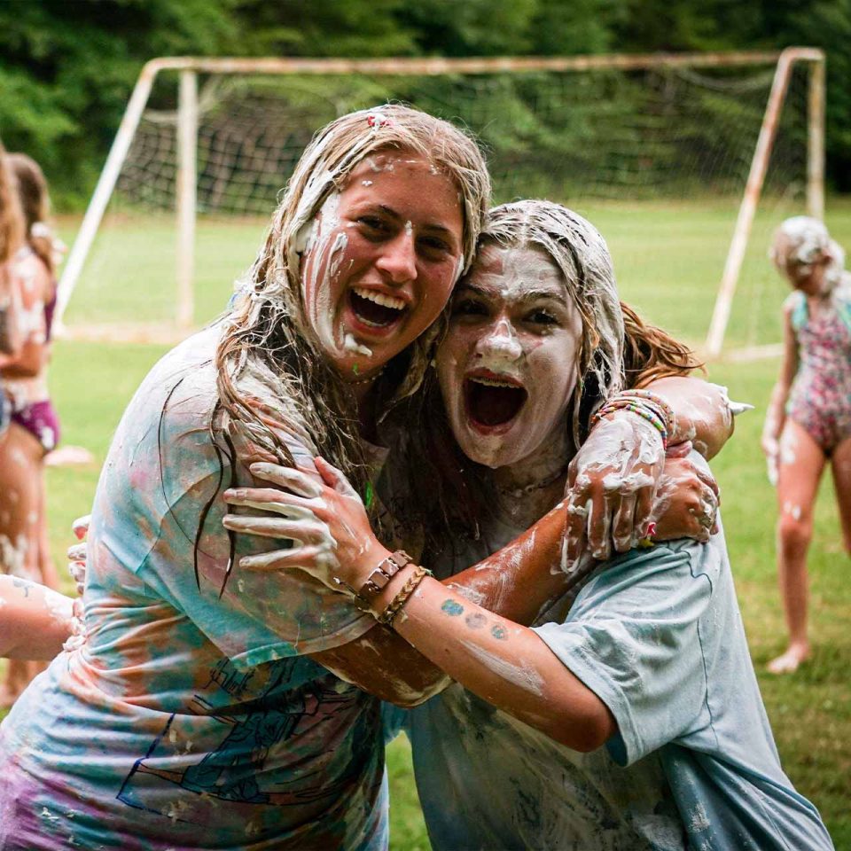 female counselor and camper embrace covered in shaving cream after a sludge war on the sports fields at Camp Canaan