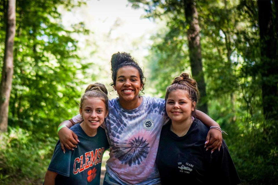 three girls embrace while smiling for the camera in nature at Camp Canaan