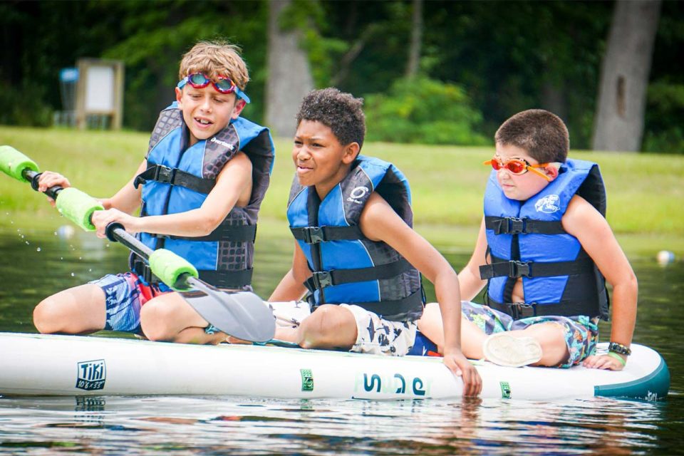 three senior day campers ride on stand up paddle board in blue life vests