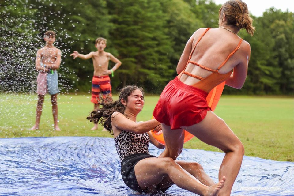 female counselors battle for inflated object while sliding on wet blue tarp in sports field at Camp Canaan