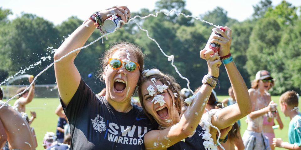 Two female counselors outdoors at Camp Canaan enthusiastically spraying each other with shaving cream, both wearing t-shirts. One person, wearing sunglasses, has "SWU Warriors" written on their shirt. Both have shaving cream on their faces and clothing, with others in the background participating in this summer camp fun.