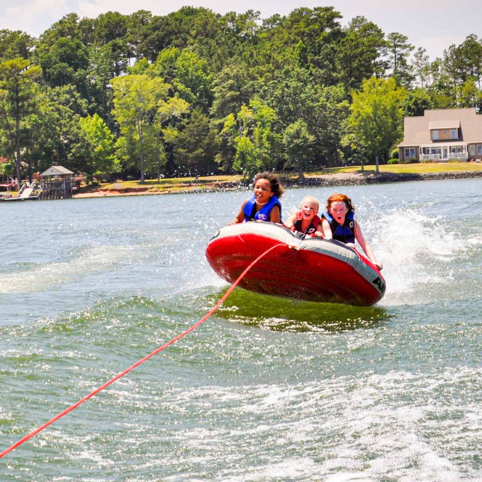 three campers hang on to red tube while being pulled behind the boat during summer camp