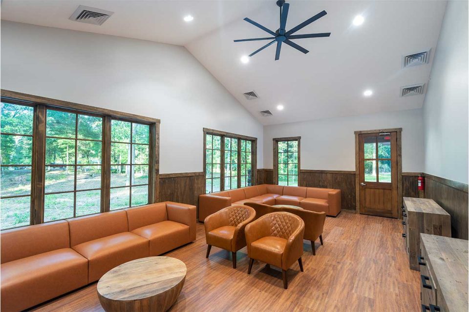 View of A&J Lodge spacious, modern common area features brown leather sofas and armchairs arranged around wooden coffee tables. Large windows provide a view of the greenery outside. Wood paneling and flooring enhance the warm, inviting atmosphere. An overhead fan hangs from the ceiling.