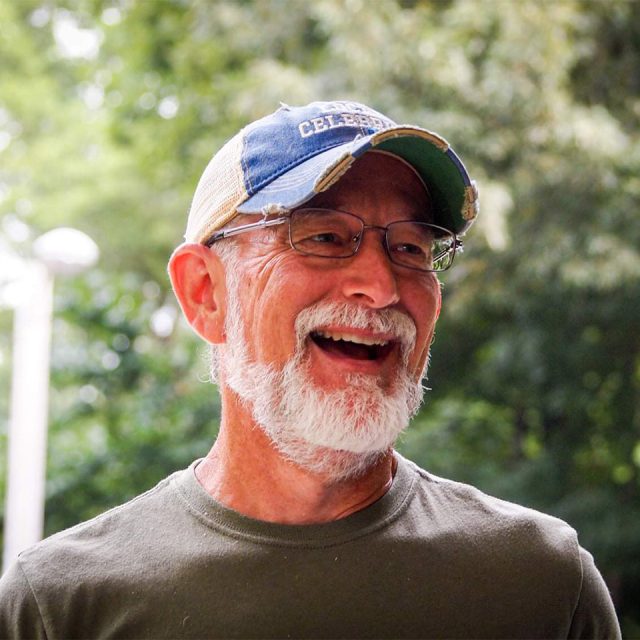 A man with a white beard and glasses is laughing outdoors at Camp Canaan. Wearing a distressed blue and beige cap and an olive green t-shirt, he stands against the blurred greenery.