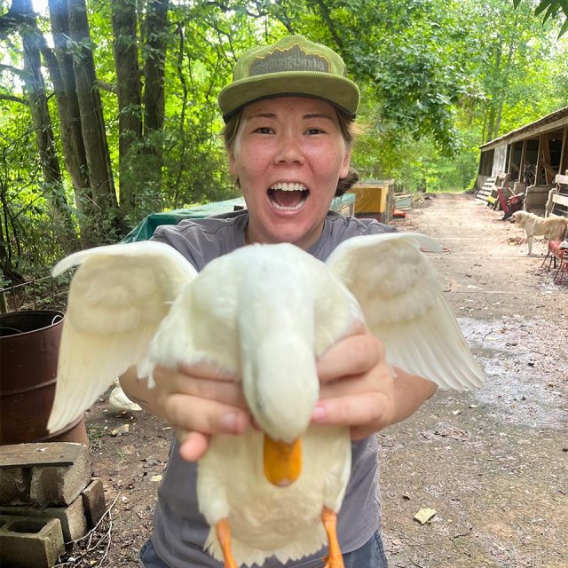 A woman wearing a green cap and gray shirt proudly holds a white duck with wings spread in front of them. They are outdoors in a wooded area with various rustic structures and equipment in the background.