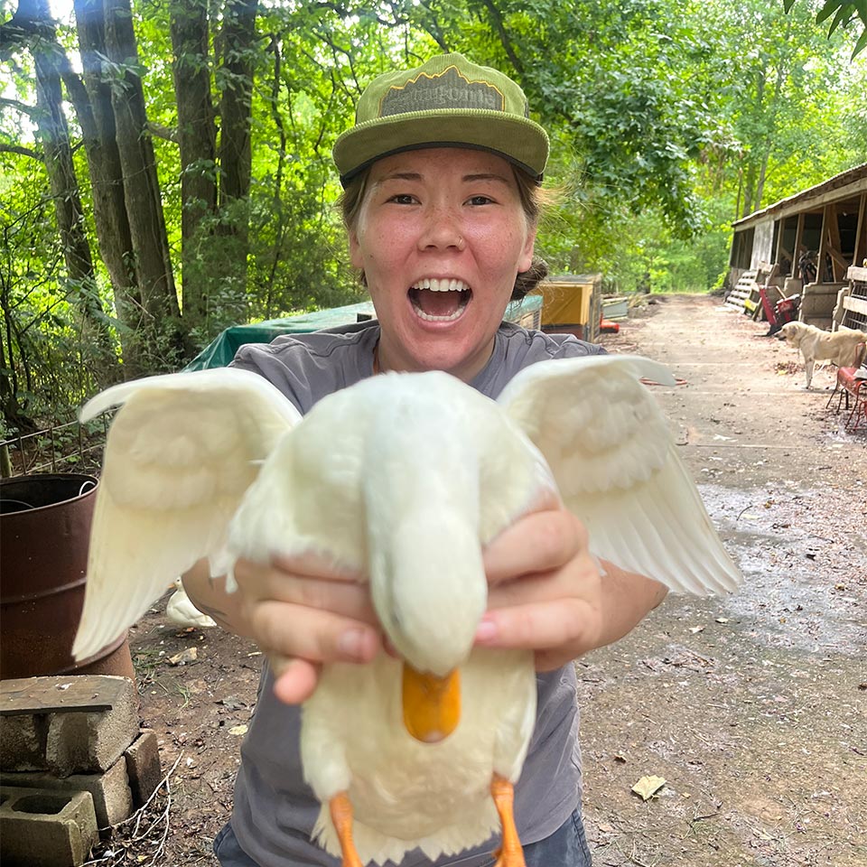 A woman wearing a green cap and gray shirt proudly holds a white duck with wings spread in front of them. They are outdoors in a wooded area with various rustic structures and equipment in the background.