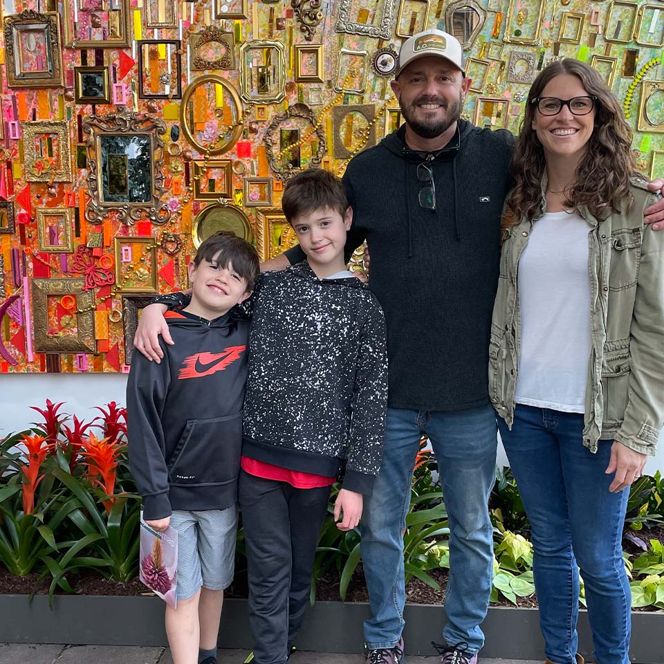 A group of four people, two adults and two boys, stand smiling in front of a colorful, framed art display.