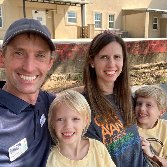 A family of four, including a man, a woman, and two young children, smiles at the camera while sitting outside. The man wears a cap and a blue shirt with a name tag. The woman and children wear casual shirts. A beige building and a wall with red paint are visible in the background.