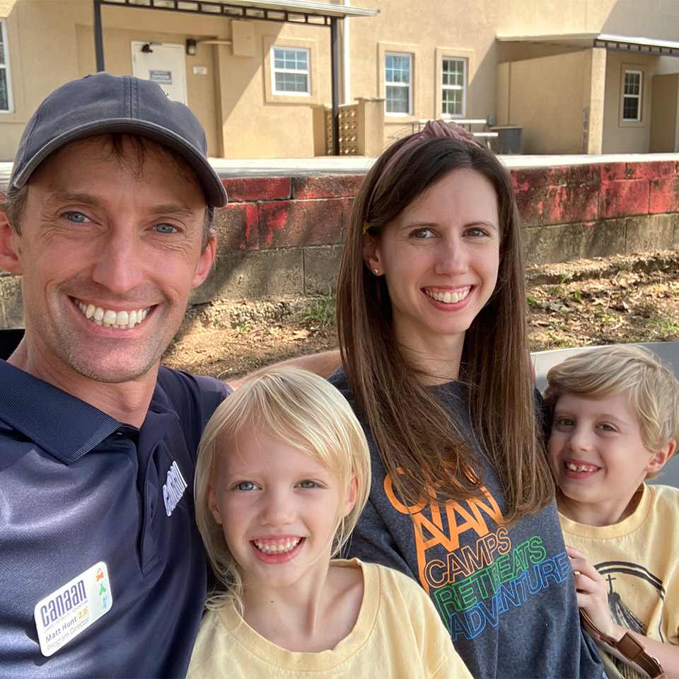 A family of four, including a man, a woman, and two young children, smiles at the camera while sitting outside. The man wears a cap and a blue shirt with a name tag. The woman and children wear casual shirts. A beige building and a wall with red paint are visible in the background.