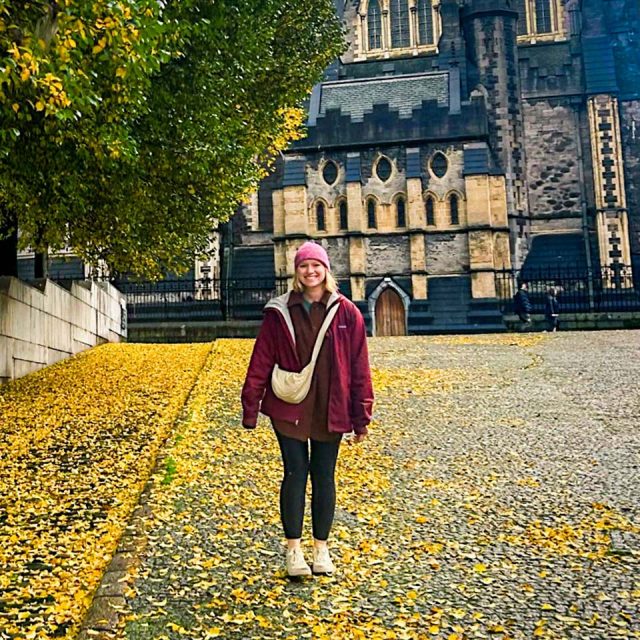 A woman wearing a maroon jacket, pink beanie, and black leggings stands under a tree with yellow leaves, with more yellow leaves scattered on the ground. In the background, there is a large stone building with Gothic architecture.