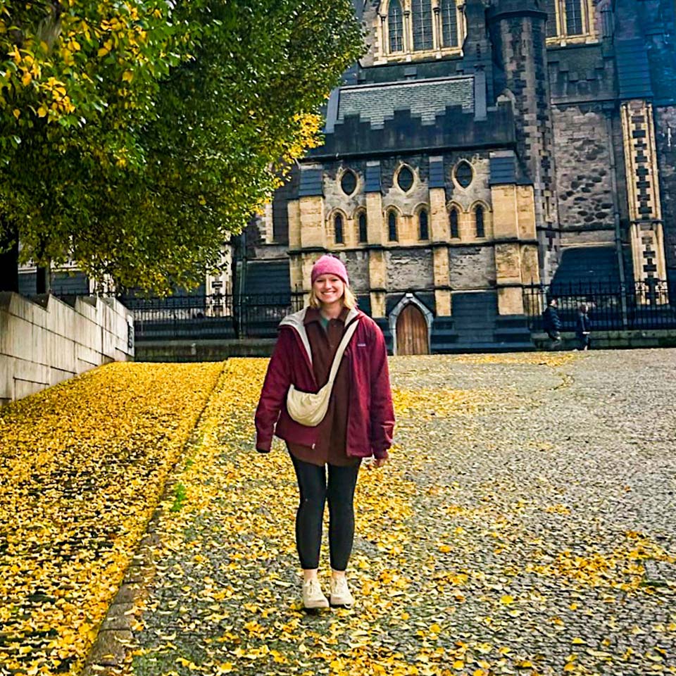 A woman wearing a maroon jacket, pink beanie, and black leggings stands under a tree with yellow leaves, with more yellow leaves scattered on the ground. In the background, there is a large stone building with Gothic architecture.