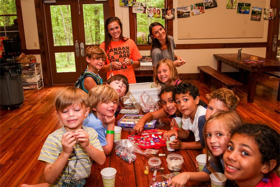 A group of children and two adults smile at the camera while sitting at a wooden table filled with arts and crafts supplies. The Multi-purpose building has wooden floors and walls, and the background shows a door leading to a green outdoor area.