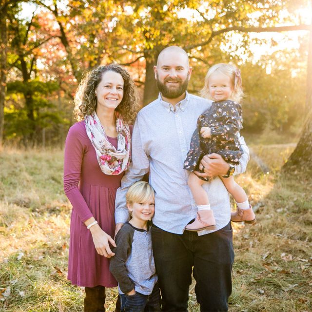 A family of four poses outdoors in an autumn setting. A woman with curly hair stands beside a bearded man holding a young girl. A young boy stands in front. Sunlight filters through the trees in the background, casting a warm glow behind them.