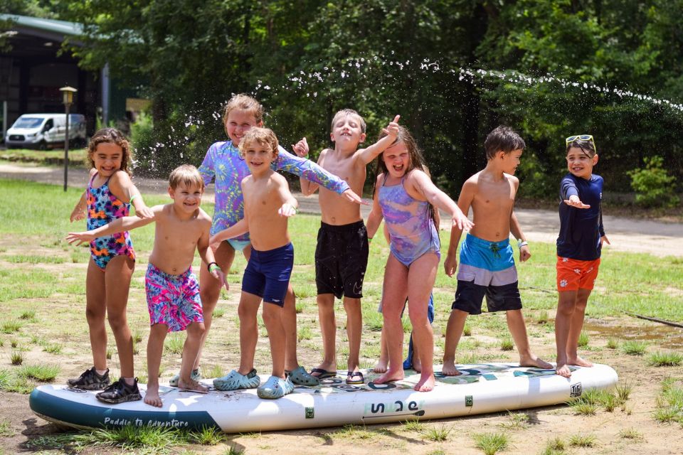 group of junior day campers pretend to surf on stand up paddle board laying on land next to Lake Canaan