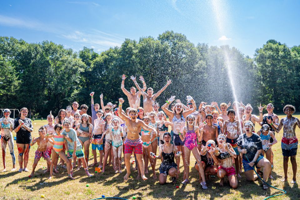 large group of campers smile for the camera in the sports field while counselor shoots water from a hose over their heads