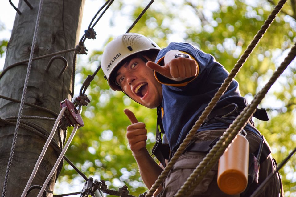 A person wearing a safety helmet and harness shows a joyful expression and gives two thumbs up while participating in an outdoor ropes course at Camp Canaan. The background shows blurred green foliage and other ropes and harness equipment.