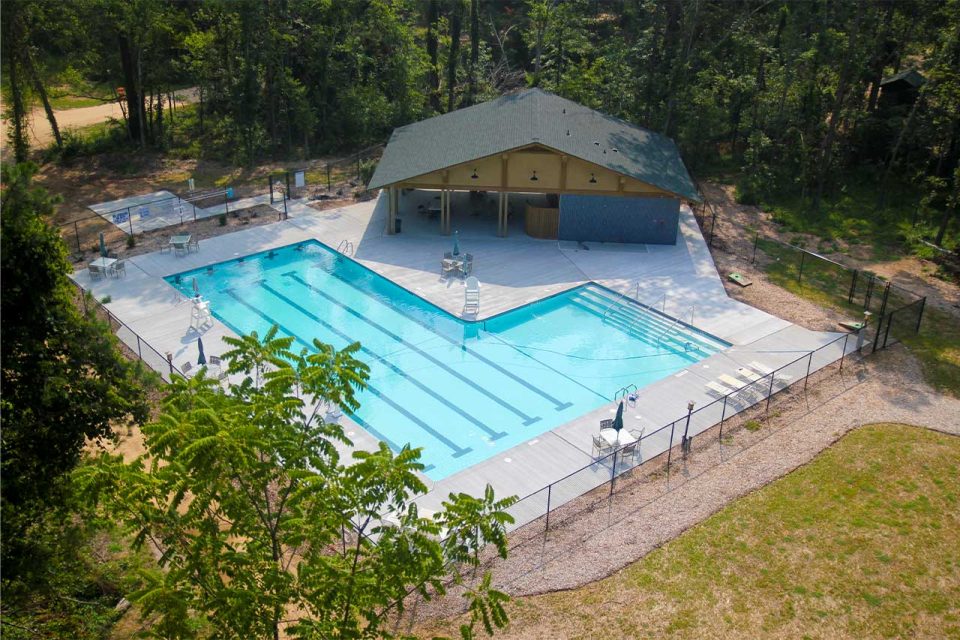 Aerial view of the swimming pool with clear blue water at Camp Canaan, surrounded by a concrete deck. The pool area includes several lounge chairs, tables with umbrellas, and a small building. Enclosed by a fence and surrounded by trees, it's perfect for an overnight summer camp experience.