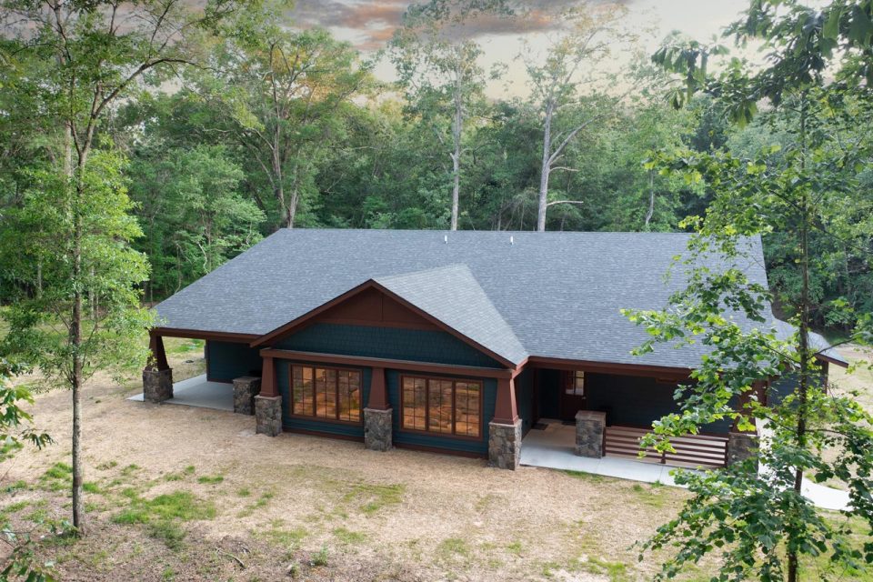 The single-story A&J Lodge at Camp Canaan has a dark exterior and a pitched roof, surrounded by tall trees. The front features large windows and stone pillars —perfect for an corporate retreat or overnight summer camp adventure.