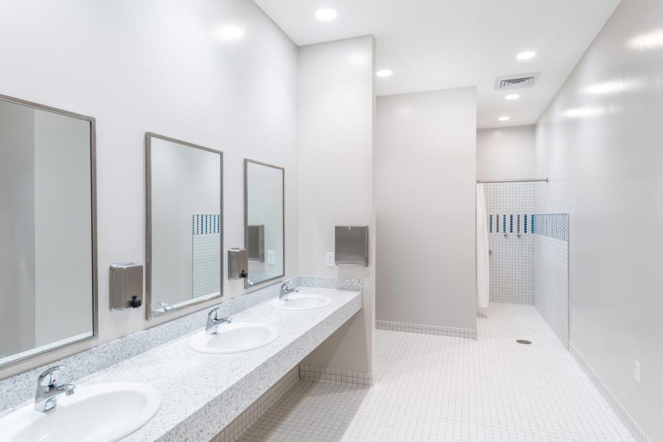 A modern restroom with a light color scheme, perfect for a retreat or overnight camp, featuring three white sinks with mirrors above each, a paper towel dispenser mounted on the wall, and a spacious tiled walk-in shower area at the back. The room is well-lit with ceiling lights.