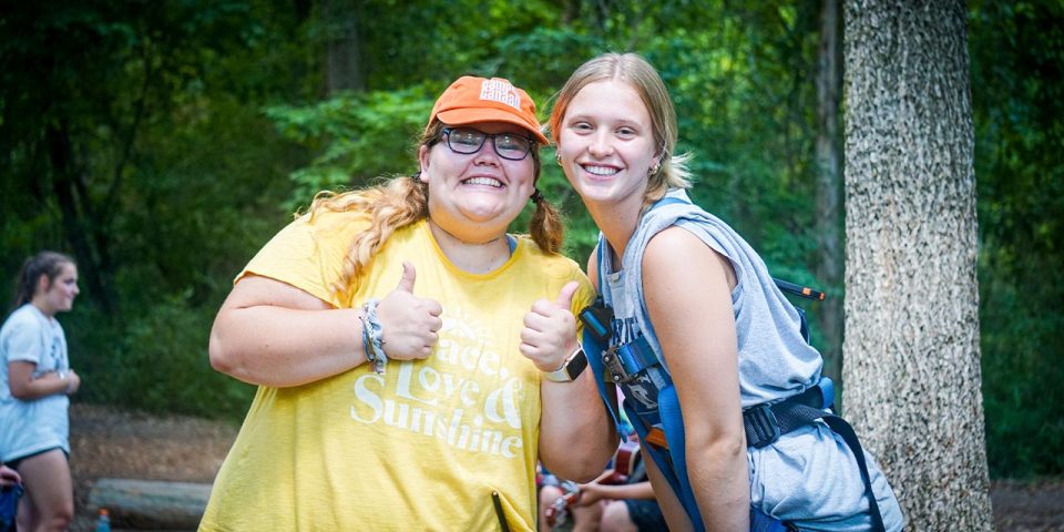 Two people smiling outdoors, one wearing a yellow shirt and an orange cap giving two thumbs up, and the other in a gray shirt with a harness. They're at Camp Canaan enjoying their summer camp experience, with trees in the background.