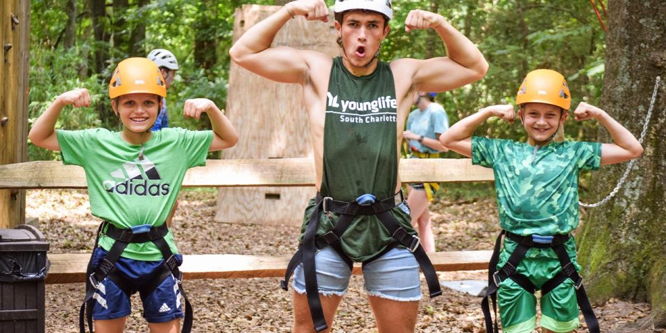 One counselor and two campers wearing helmets and harnesses stand in front of a wooden fence in a wooded area at Camp Canaan. The person in the middle flexes their arms and makes an expressive face, while the two children on either side mirror the flexing pose and smile.