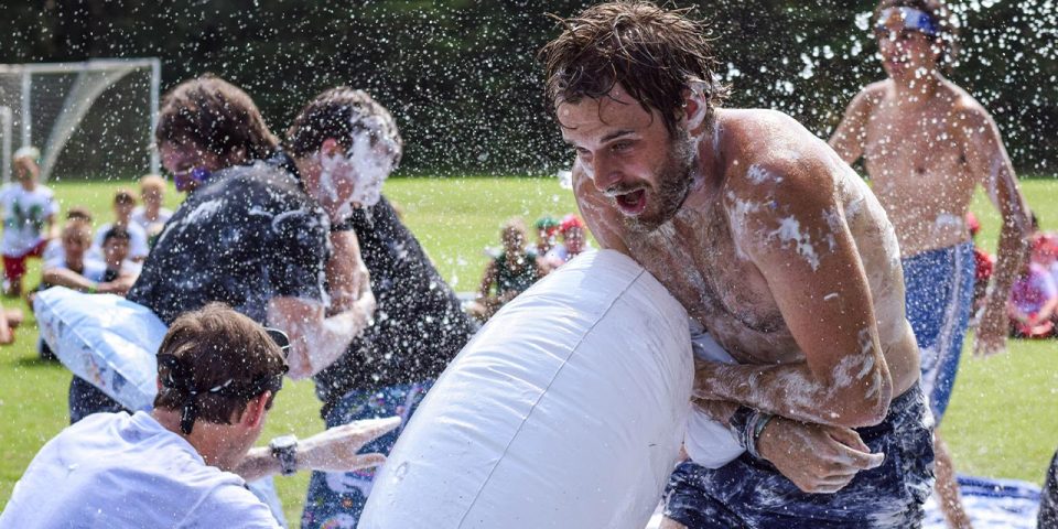 A group of people at a summer camp are playing an outdoor game on the sports field. One person in the foreground clutches an inflatable pool tool covered in soap suds and water, while others engage in the background.