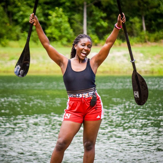 A counselor in red shorts and a black tank top stands joyfully in front of Lake Canaan, holding paddles high in each hand. The backdrop is a serene lake surrounded by lush greenery.