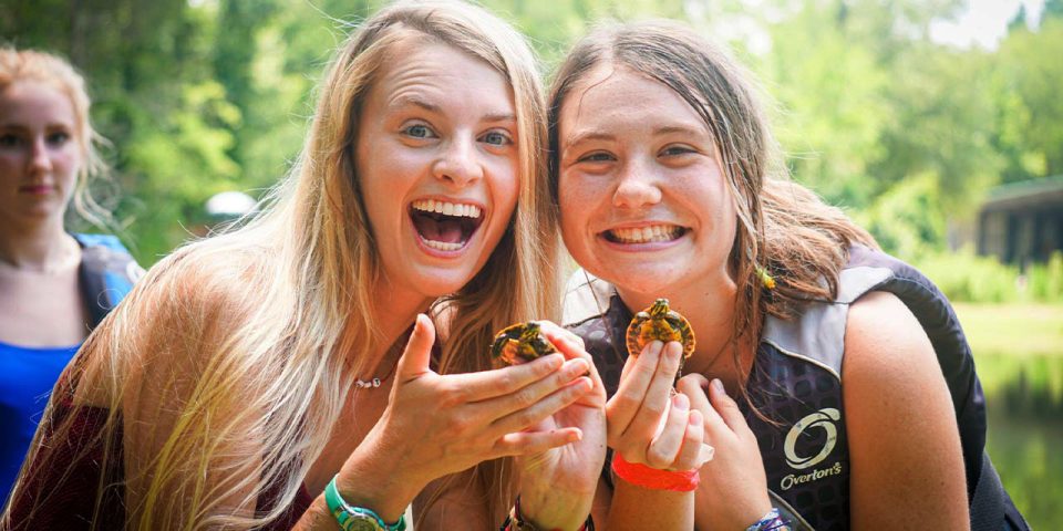 Two smiling counselors hold baby turtles in their hands during overnight summer camp. They are outdoors, with trees and a body of water in the background.