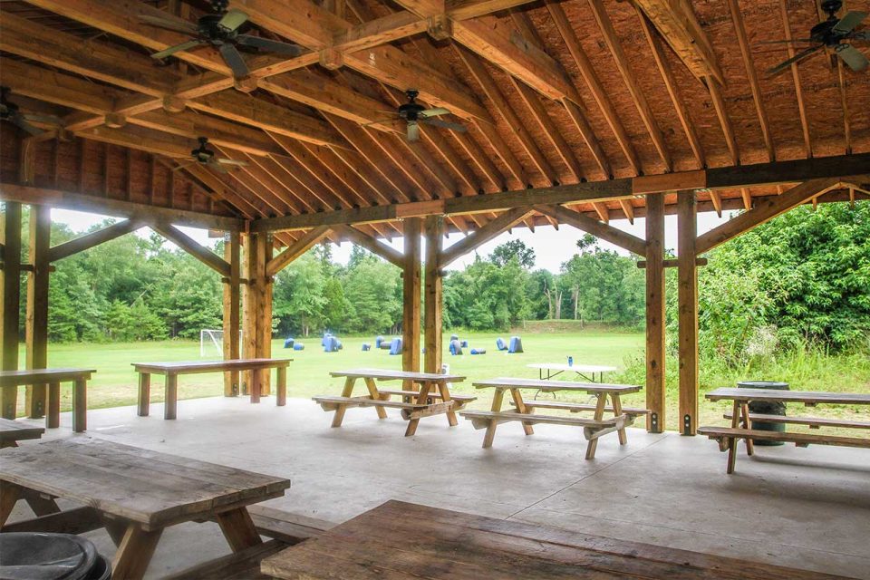 Picnic pavilion with wooden beams and picnic tables underneath. The area overlooks a grassy field.