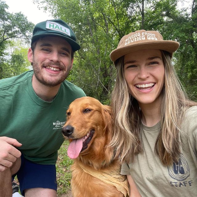 Two people smiling, one man and one woman, are posing with a golden retriever outdoors at Camp Canaan. They are wearing casual clothes and caps, with trees and greenery in the background.