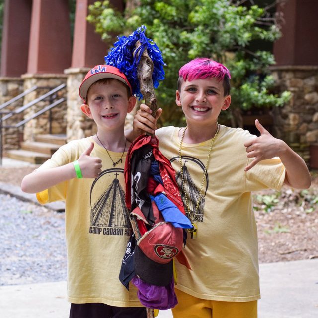 Two kids smile and give thumbs-up gestures while standing close together outside the multi-purpose building. They wear matching yellow Camp Canaan t-shirts with beaded necklaces.