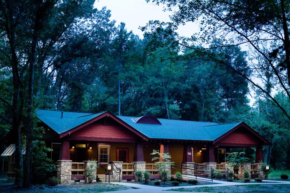 A rustic cabin with a pitched roof is nestled among tall trees at Camp Canaan. The single-story structure features a stone foundation, wooden siding, and a oversized porch. Soft lighting illuminates the entrance, giving a warm, inviting atmosphere perfect for summer camp activities or corporate retreats.