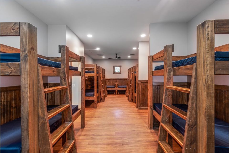 View of A&J Lodge with wooden bunk beds arranged in two rows along the walls. Each bunk bed has a ladder, and the lower bunks are enclosed with wooden paneling. The room has wood-style flooring and bright recessed lighting. A small window is visible at the far end of the room.