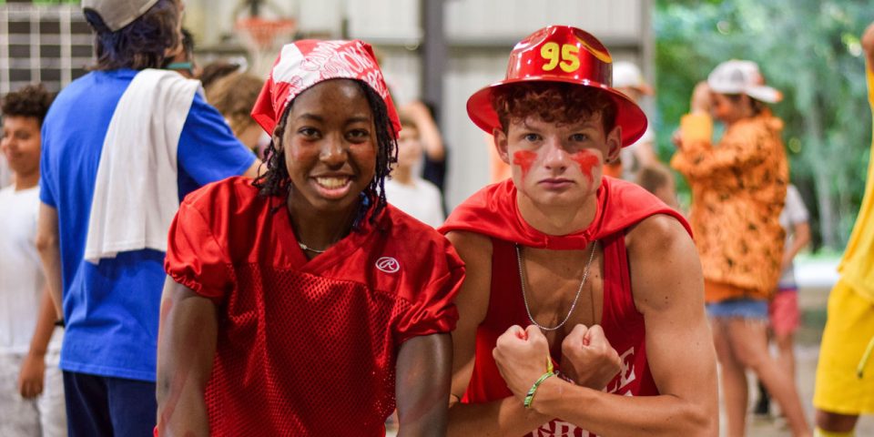 Two people in colorful costumes pose for a photo during Champion's week at Camp Canaan. The person on the left, smiling, wears a red bandana, red top, and shorts. The person on the right wears a red hard hat with number 95, red cape, and war paint, striking a superhero pose.