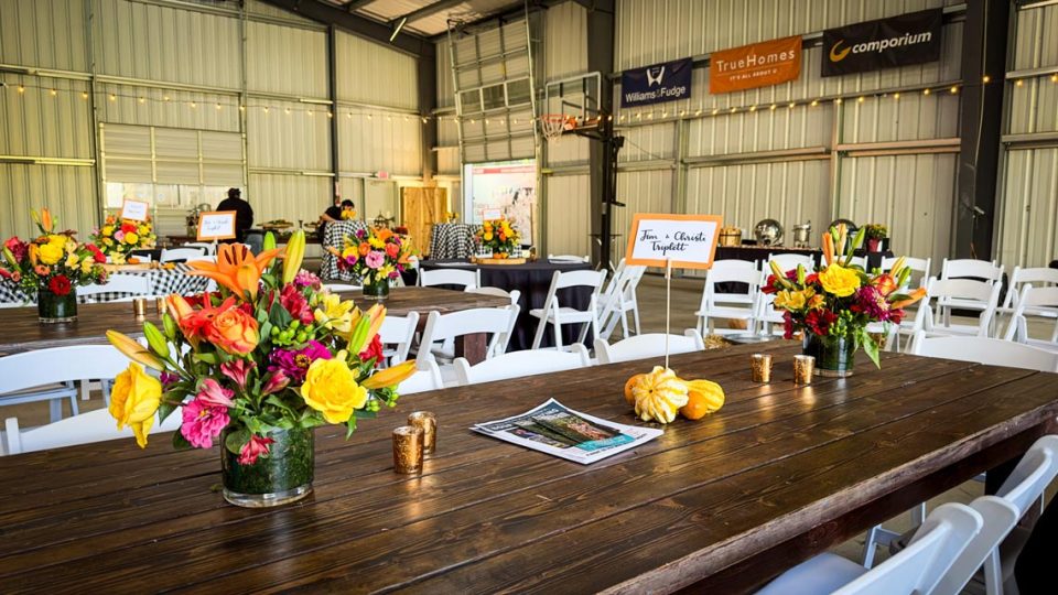 A decorated event space inside the covered basketball court with wooden tables set up with floral centerpieces and small pumpkins. Company banners for Camp Canaan sponsors are visible on the wall. White folding chairs surround the tables.