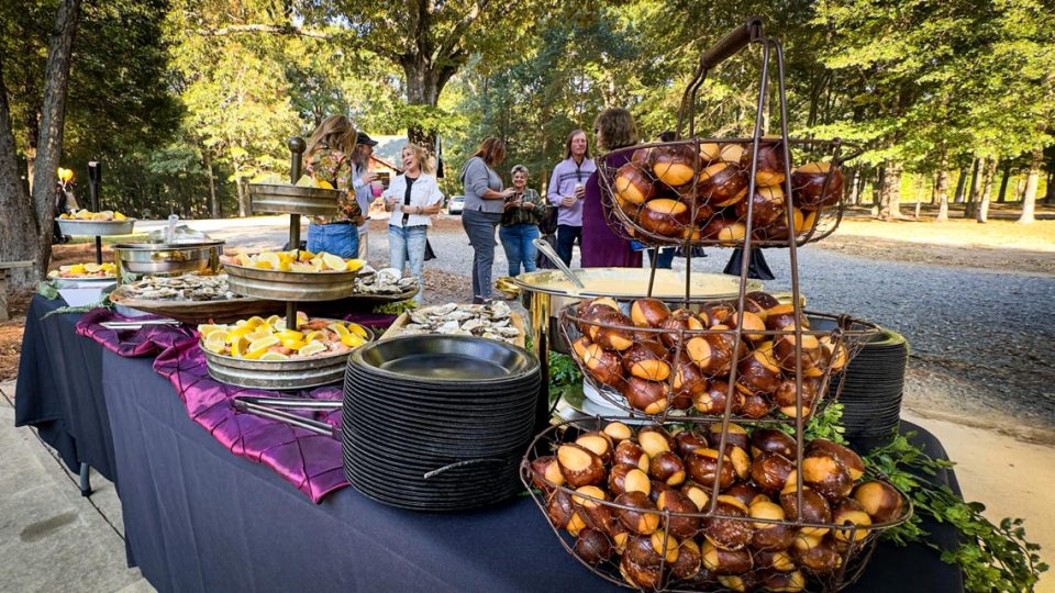 An outdoor buffet table at Camp Canaan is set up with a variety of food, including pretzel bitesin a tiered basket, shrimp, and other dishes. A group of people stands nearby, engaged in conversation. The setting is surrounded by trees and greenery, suggesting a casual event.