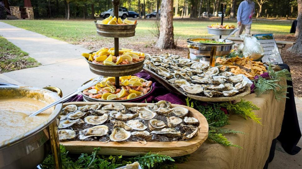 A buffet table set outdoors at the retreat center features a variety of seafood. A wooden tray filled with oysters, a tiered stand of sliced lemons and shrimp, and multiple bowls containing other food items invite guests to indulge. In the background, trees and a pathway are visible.