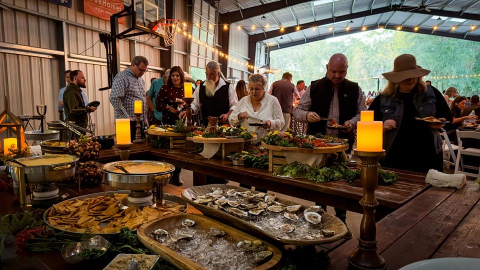 A group of people at Camp Canaan are serving themselves food from a buffet table inside the covered basketball court. The table is adorned with various dishes including oysters, pretzel bites, and dips. The space is warmly lit with decorative lights and candles, all set against a backdrop of trees.