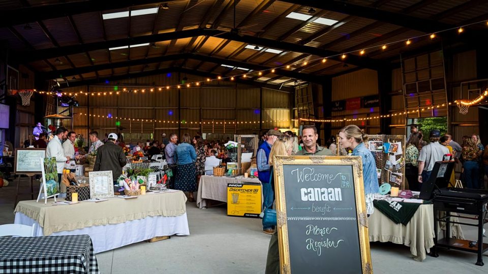 A busy indoor event features people mingling and browsing various tables filled with silent auction items. String lights decorate the ceiling, creating a festive atmosphere.
