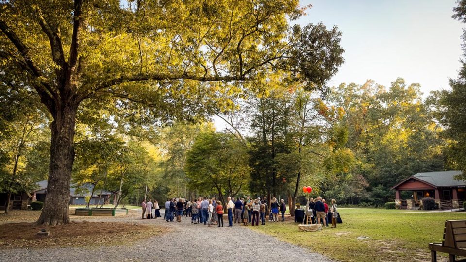 A group of people gather outdoors under a large tree at Camp Canaan for the annual Canaan benefit. They are standing around tables covered with checkered cloths, engaged in conversation.