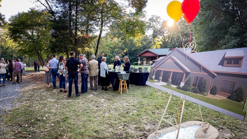 People attending the outdoor event with trees in the background are mingling near tables with food and drinks. A large poster of a future Camp Canaan dining hall is displayed on an easel with two helium balloons, one red and one yellow, tied to it.