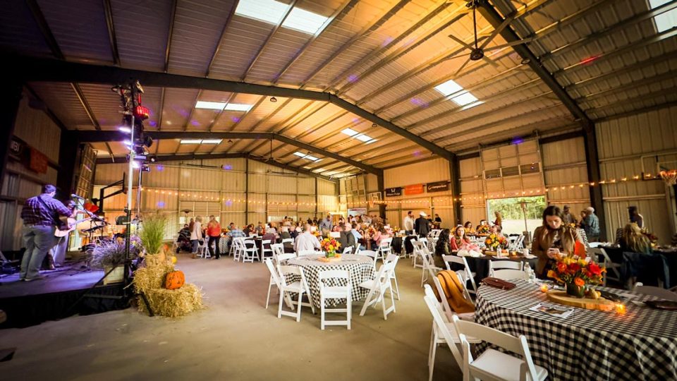 People seated at round tables covered in checkered tablecloths in the covered basketball court. String lights decorate the space. A stage with musicians is on the left, and various fall-themed decorations, including pumpkins, are visible.
