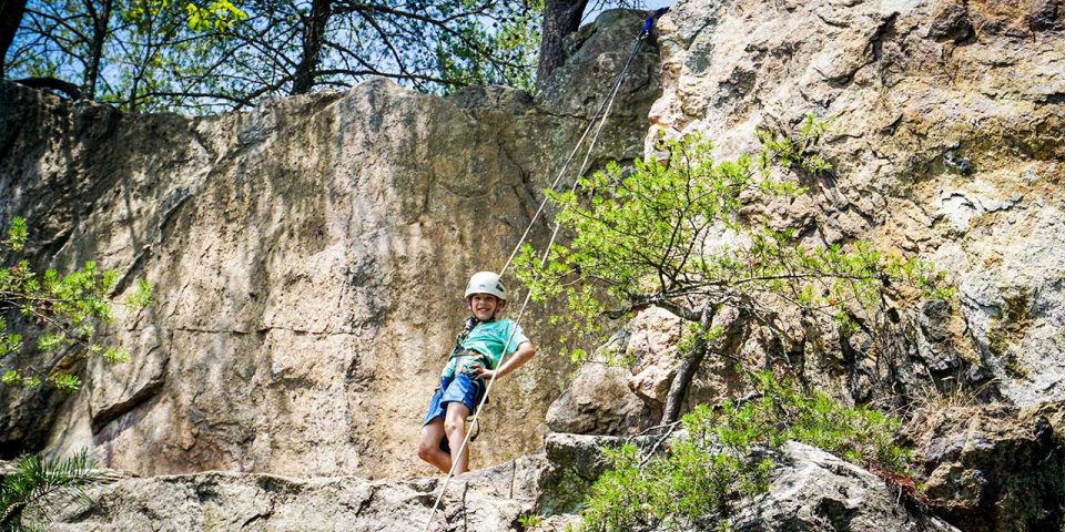 A young camper wearing a helmet climbs a steep rock face, holding onto a rope. The child is at Crowders Mountain, surrounded by large rocky formations and sparse greenery.