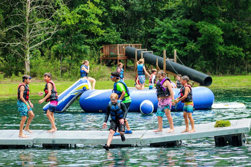 Children wearing life jackets are playing on and around a floating dock and large inflatable water structures in the lake at Camp Canaan. Trees and a large slide are in the background, capturing the essence of summer camp.