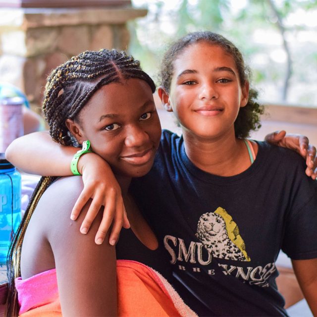 Two smiling girls sit closely together at Camp Canaan with their arms around each other. One wears a strapless top, the other a T-shirt. In the background, another person is eating a popsicle and holding a water bottle.